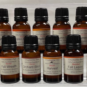 Op de afbeelding: Een verzameling amberkleurige glazen etherische olieflessen met zwarte doppen. Elke fles heeft een etiket met de merknaam en geurbeschrijving, waaronder "Spiced Cider", "Pumpkin Pie" en "Fall Spice". De oliën zijn gemengd.