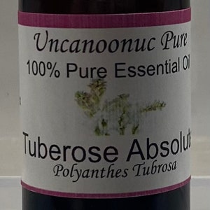 May include: A dark amber glass bottle with a black cap, labeled "Uncanoonuc Pure 100% Pure Essential Oil". The label also reads "Tuberose Absolute, Polianthes Tubrosa". The label has a pink border and a floral illustration.