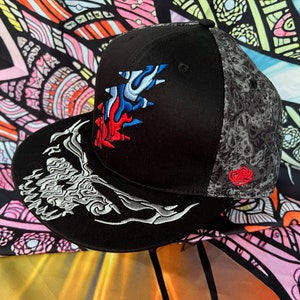 May include: Black baseball cap with a grey and white skull design on the brim and a colorful embroidered graphic on the front. The graphic features a red, blue, and white tree-like design.