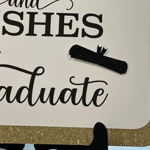 Peut inclure: Une pancarte blanche avec une bordure dor&eacute;e scintillante et une inscription noire qui dit "and wishes the graduate". Un dipl&ocirc;me noir scintillant est attach&eacute; &agrave; la pancarte.