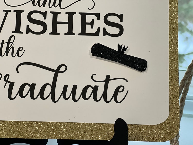 Peut inclure: Une pancarte blanche avec une bordure dor&eacute;e scintillante et une inscription noire qui dit "and wishes the graduate". Un dipl&ocirc;me noir scintillant est attach&eacute; &agrave; la pancarte.
