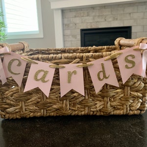 May include: A wicker basket with a pink and gold banner that reads "Cards". The banner is strung across the top of the basket and has gold glitter letters.