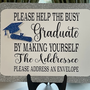 May include: A white sign with black text that reads "Please help the busy graduate by making yourself the addressee. Please address an envelope." The sign has a blue glitter graduation cap and diploma on it.