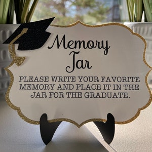 May include: A white sign with gold glitter trim and a black graduation cap graphic. The sign reads "Memory Jar" and "Please write your favorite memory and place it in the jar for the graduate."