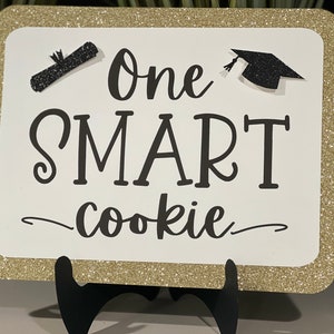 May include: A white sign with gold glitter trim and the text "One SMART cookie" in black. The sign has a graduation cap and diploma graphic. The sign is displayed on a black easel.