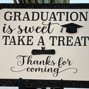 May include: Black and white sign with the text "Graduation is sweet take a treat" and "Thanks for coming". The sign has a graduation cap and diploma graphic.