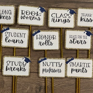 May include: Nine gold glitter graduation signs with blue graduation caps. The signs have various sayings including "So Long Suckers", "Book Worms", "Class Rings", "Thank You Kisses", "Student Loans", "Honor Rolls", "Reach for the Stars", "Study Breaks", "Nutty Professors", and "Smartie Pants".