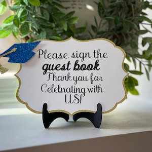 May include: White sign with gold glitter trim and a blue graduation cap on top. The sign reads "Please sign the guest book Thank you for celebrating with US!"