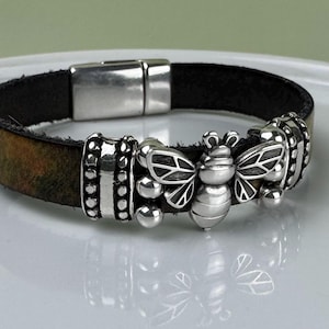 Puede incluir: Una pulsera de cuero con un cierre magnético plateado y abalorios decorativos de abeja plateados. La pulsera es de color marrón oscuro y negro, con un acabado envejecido. Los abalorios de abeja tienen alas detalladas y un cuerpo redondeado.