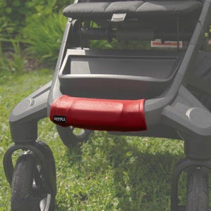 Può includere: Una copertura per il manubrio del passeggino in pelle rossa. La copertura è rettangolare e avvolge il manubrio del passeggino. La parola "SADDYALE" è stampata sulla copertura. Il passeggino è grigio e nero, con un sedile e ruote neri. Lo sfondo è erba verde.