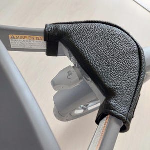 以下が含まれることがあります： 黒い革製のベビーカーのハンドルカバー。カバーは質感があり、ベビーカーのハンドルを包み込みます。ベビーカーのフレームはグレーで、警告ラベルが見えます。