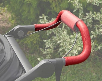 Può includere: Primo piano di un manubrio per passeggino in pelle rossa. Il manubrio è curvo e ha una superficie strutturata. Il telaio in metallo è grigio. Lo sfondo è sfocato con fogliame verde.