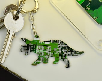 May include: A silver key chain with three keys and a green circuit board dinosaur keychain. The dinosaur keychain is made from recycled circuit boards.