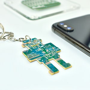 May include: A green and gold circuit board keychain shaped like a robot. The keychain is attached to a silver key ring with a gold key.