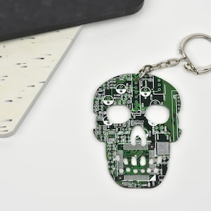 May include: A green circuit board shaped like a skull with a silver key chain attached. The skull has a detailed design of a circuit board with white and black accents.
