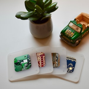 May include: Four white square coasters with different circuit board designs. The coasters are on a white surface next to a small green toy jeep with a tan top and the number "337" on the side.
