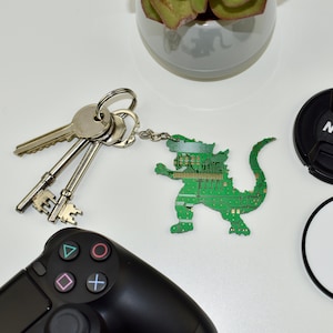 May include: A green circuit board keychain shaped like a monster, a set of keys, a black and white video game controller, a black lens cap with the word "Nikon" printed on it, and a white round sticker with a black logo.