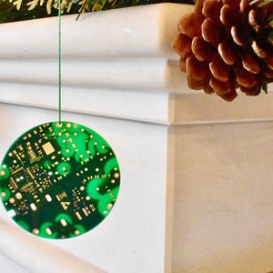 May include: A green and gold circuit board Christmas ornament hanging from a green string. The ornament is round and has a detailed design.