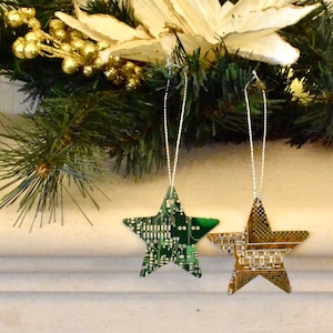Może przedstawiać: Dwie zielono-złote, gwiazdkowe ozdoby choinkowe wykonane z recyklingowanych płytek drukowanych. Ozdoby wiszą na srebrnych sznurkach.