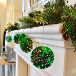 May include: Green Christmas ornaments shaped like circuit boards hang from a white fireplace mantel. The ornaments are decorated with gold accents.