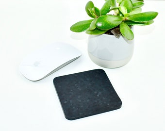 May include: A black square mouse pad with a speckled texture sits on a white surface next to a white wireless computer mouse and a small potted succulent plant.
