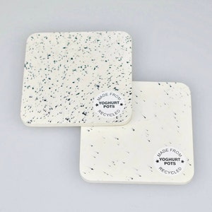 Op de afbeelding: Twee witte vierkante onderzetters met zwarte stippen. De onderzetters zijn gemaakt van gerecyclede yoghurtbekers en hebben een zwart-witte sticker met de tekst "Gemaakt van gerecyclede yoghurtbekers".