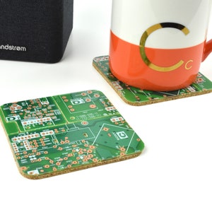 May include: Two green circuit board coasters with cork backing. The coasters feature copper traces and components. A white mug with an orange bottom and a gold letter "C" is sitting on one of the coasters.