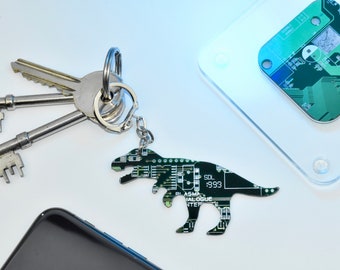 May include: A green circuit board dinosaur keychain with a silver key ring and three keys. The dinosaur is a T-Rex with a bottle opener on its side. The circuit board has the text "SDL 1993" on it.