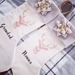 Puede incluir: Dos calcetines de lona blancos con un dise&ntilde;o de ciervo en chevron rosa y blanco. Un calcet&iacute;n dice "Granddad" y el otro dice "Nanna".