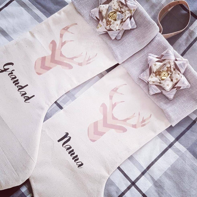 Puede incluir: Dos calcetines de lona blancos con un dise&ntilde;o de ciervo en chevron rosa y blanco. Un calcet&iacute;n dice "Granddad" y el otro dice "Nanna".
