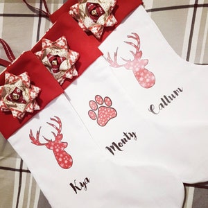 Puede incluir: Tres calcetines de Navidad blancos con pu&ntilde;os y lazos de fieltro rojo. Los calcetines est&aacute;n personalizados con los nombres "Kya", "Monty" y "Callum". Cada calcet&iacute;n presenta un dise&ntilde;o de reno rojo o una huella de pata.