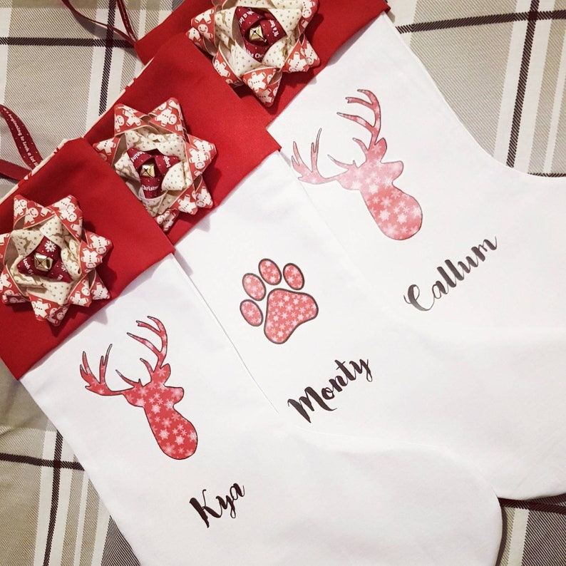Puede incluir: Tres calcetines de Navidad blancos con pu&ntilde;os y lazos de fieltro rojo. Los calcetines est&aacute;n personalizados con los nombres "Kya", "Monty" y "Callum". Cada calcet&iacute;n presenta un dise&ntilde;o de reno rojo o una huella de pata.