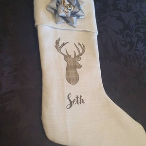 Puede incluir: Calcet&iacute;n de Navidad blanco con una silueta de ciervo marr&oacute;n y un patr&oacute;n de cuadros. El calcet&iacute;n tiene un lazo plateado y blanco y el nombre "Seth" est&aacute; bordado en la parte delantera.