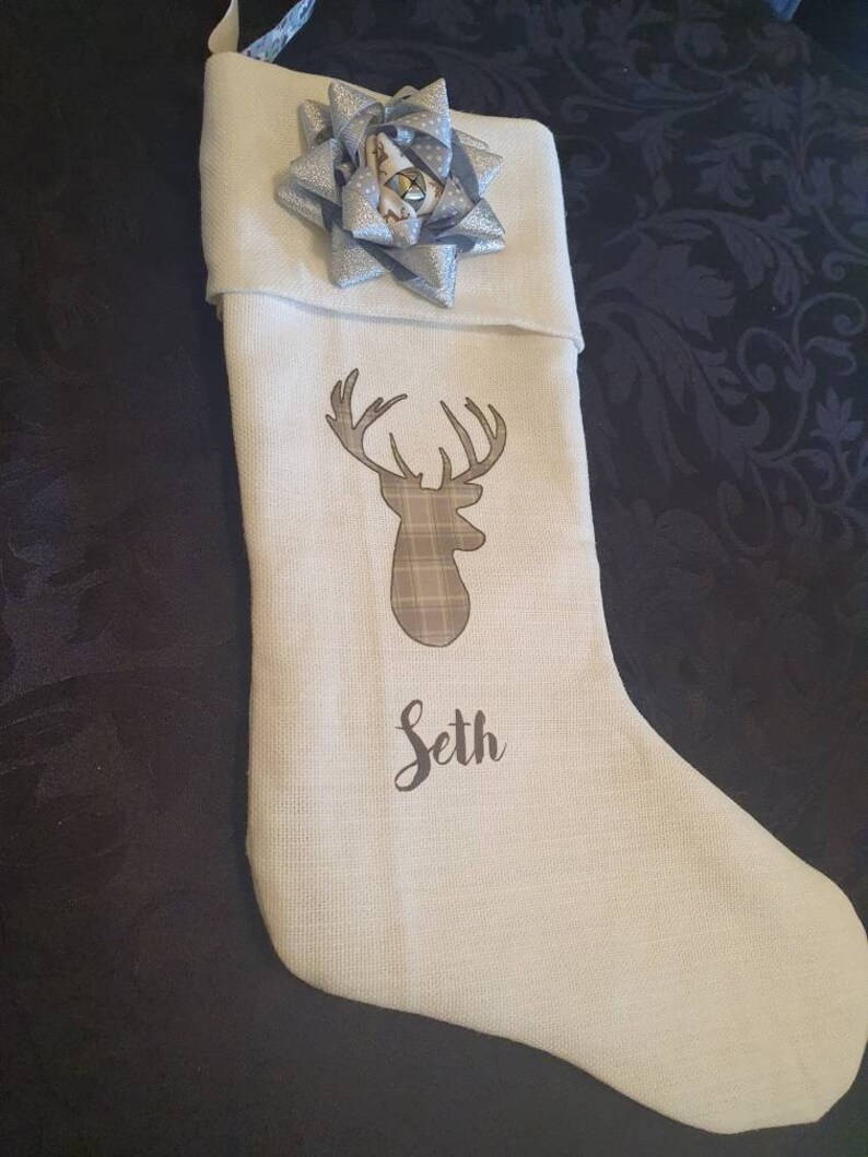 Puede incluir: Calcet&iacute;n de Navidad blanco con una silueta de ciervo marr&oacute;n y un patr&oacute;n de cuadros. El calcet&iacute;n tiene un lazo plateado y blanco y el nombre "Seth" est&aacute; bordado en la parte delantera.