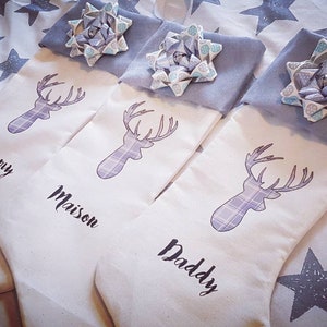 Puede incluir: Tres calcetines de Navidad blancos con un dise&ntilde;o de ciervo de cuadros grises y un lazo azul en la parte superior. Los calcetines est&aacute;n personalizados con los nombres "Mummy", "Maison" y "Daddy".