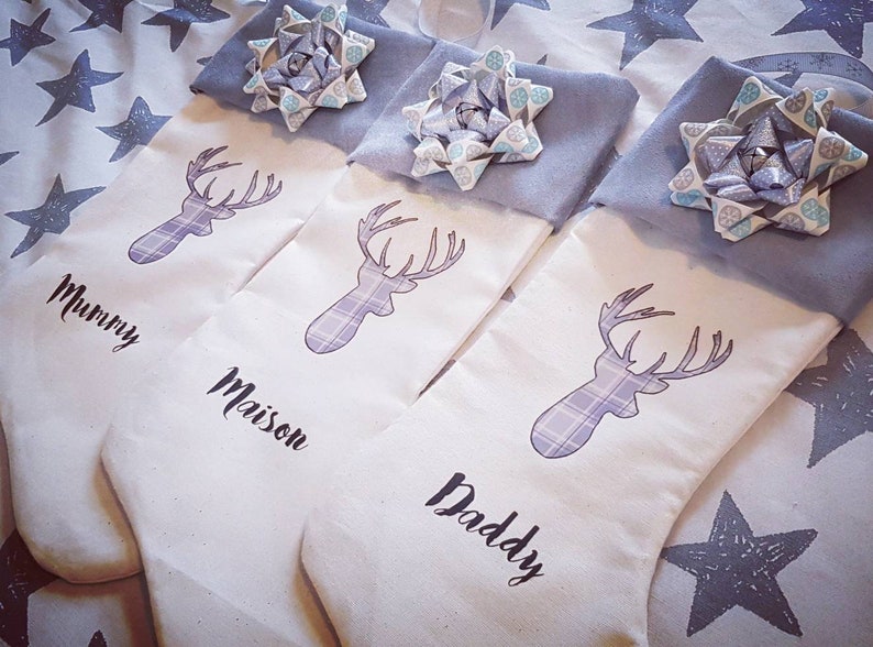 Puede incluir: Tres calcetines de Navidad blancos con un dise&ntilde;o de ciervo de cuadros grises y un lazo azul en la parte superior. Los calcetines est&aacute;n personalizados con los nombres "Mummy", "Maison" y "Daddy".