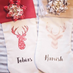 Puede incluir: Dos calcetines de Navidad blancos con dise&ntilde;os de renos rojos y rosas. Los calcetines est&aacute;n personalizados con los nombres "Tobias" y "Hamish". Cada calcet&iacute;n tiene un gran lazo en la parte superior.