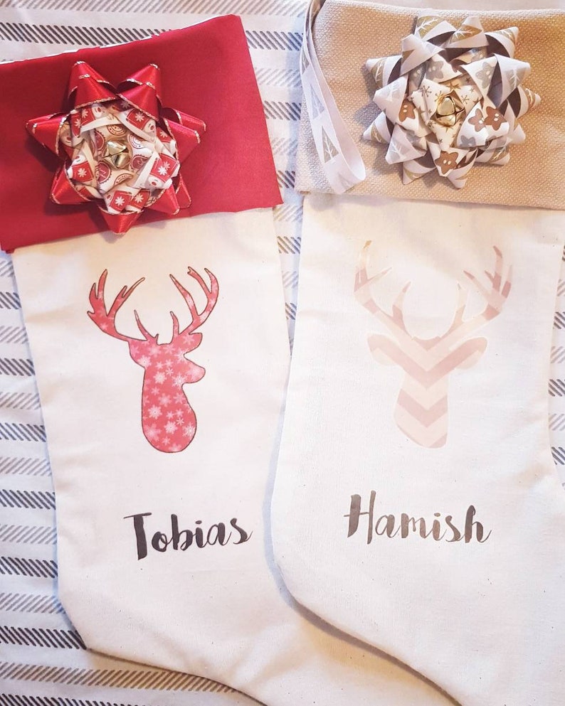 Puede incluir: Dos calcetines de Navidad blancos con dise&ntilde;os de renos rojos y rosas. Los calcetines est&aacute;n personalizados con los nombres "Tobias" y "Hamish". Cada calcet&iacute;n tiene un gran lazo en la parte superior.