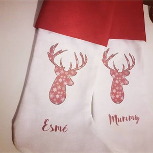 Puede incluir: Dos calcetines de Navidad blancos con pu&ntilde;os de fieltro rojo. Cada calcet&iacute;n presenta un reno rosa con un patr&oacute;n de copo de nieve y un nombre personalizado. El calcet&iacute;n de la izquierda dice "Esme" y el calcet&iacute;n de la derecha dice "Mummy".