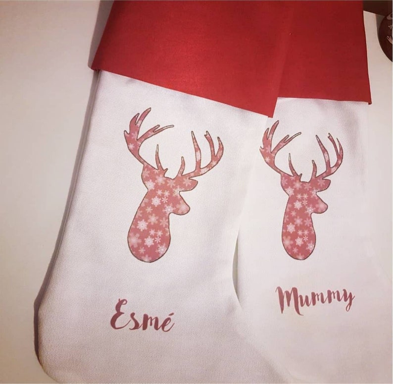 Puede incluir: Dos calcetines de Navidad blancos con pu&ntilde;os de fieltro rojo. Cada calcet&iacute;n presenta un reno rosa con un patr&oacute;n de copo de nieve y un nombre personalizado. El calcet&iacute;n de la izquierda dice "Esme" y el calcet&iacute;n de la derecha dice "Mummy".