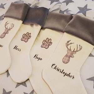 Puede incluir: Cuatro calcetines de Navidad blancos con pu&ntilde;os de terciopelo gris. Los calcetines est&aacute;n decorados con un ciervo, una huella de pata y los nombres "Catherine", "Floss", "Hugo" y "Christopher".