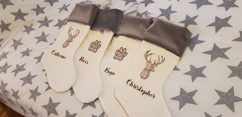 Puede incluir: Cuatro calcetines de Navidad blancos con pu&ntilde;os de terciopelo gris. Los calcetines est&aacute;n decorados con un ciervo, una huella de pata y los nombres "Catherine", "Floss", "Hugo" y "Christopher".