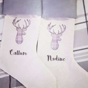 Puede incluir: Dos calcetines de Navidad blancos con pu&ntilde;os de fieltro gris. Cada calcet&iacute;n presenta un dise&ntilde;o de ciervo de cuadros morado y blanco y un nombre personalizado. Los nombres son "Callum" y "Nadine".