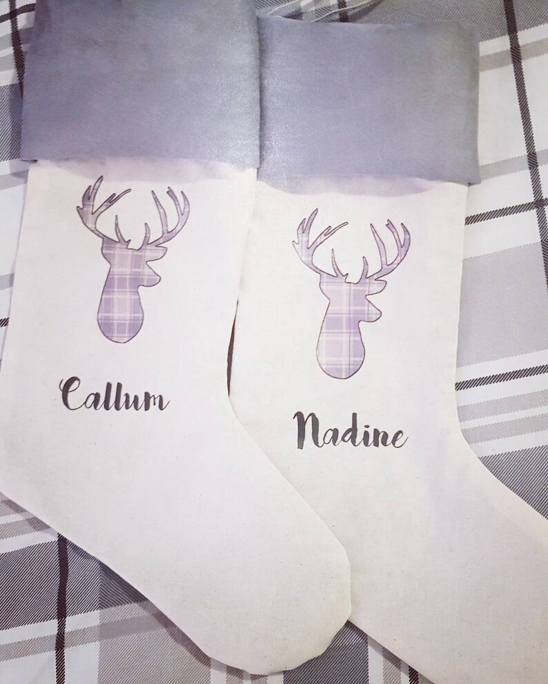 Puede incluir: Dos calcetines de Navidad blancos con pu&ntilde;os de fieltro gris. Cada calcet&iacute;n presenta un dise&ntilde;o de ciervo de cuadros morado y blanco y un nombre personalizado. Los nombres son "Callum" y "Nadine".