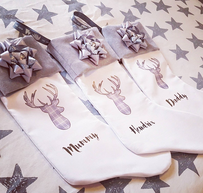 Puede incluir: Tres calcetines de Navidad blancos con un pu&ntilde;o de fieltro gris y un lazo gris. Cada calcet&iacute;n presenta un ciervo blanco con un patr&oacute;n de cuadros y un nombre: "Mummy", "Hendrix" y "Daddy".