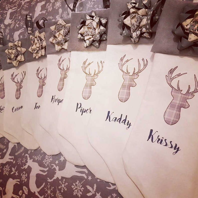 Puede incluir: Seis calcetines blancos con tapas y lazos de fieltro gris. Cada calcet&iacute;n presenta un dise&ntilde;o de cabeza de ciervo a cuadros y un nombre: 'Josi', 'Caison', 'Tor', 'Harpe', 'Piper', 'Kaddy' y 'Krissy'.