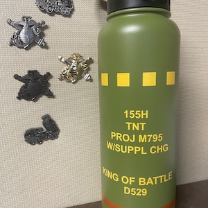 May include: An olive green water bottle with a black lid and yellow text that reads "155H TNT PROJ M795 W/SUPPL CHG KING OF BATTLE D529". Several metal pins with military-themed designs are scattered around the bottle.
