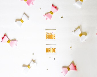 May include: White and pink bows with gold accents and two cards with the text "Team Bride" and "Bride" in orange letters on a white background.