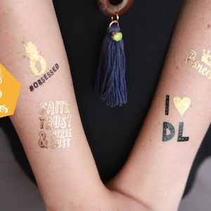 Puede incluir: Conjunto de seis tatuajes temporales dorados con varios diseños, que incluyen una piña con el texto "#obsessed", las palabras "Faith Trust & Pixie Dust", una corona con la palabra "Princess", y el texto "I love DL" con un corazón dorado.