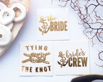 May include: Three gold temporary tattoos with nautical themes. The first tattoo says "the bride" with an anchor. The second tattoo says "tying the knot" with a rope knot. The third tattoo says "bride's crew" with an anchor.